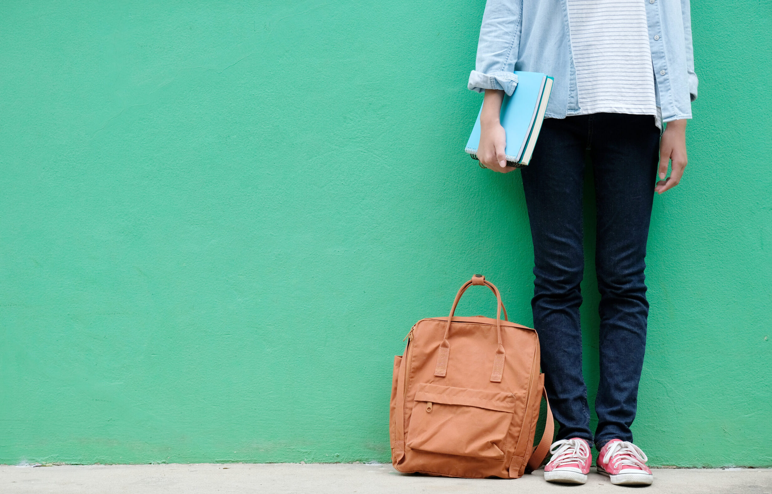 the image shows the bottom half of a young person standing and holding their books, with their bag by their feet