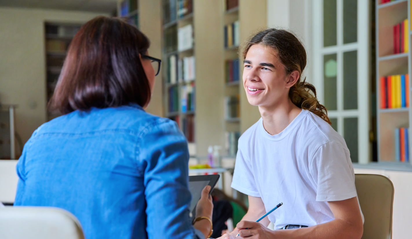 the photo shows a meeting between a female adult and a male young person, who is smiling at the adult and holding a pencil and clipboard